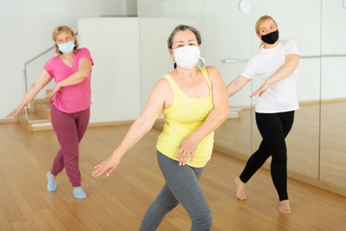 Three mature European women in masks are dancing in fitness room